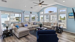 A spacious living room with a large sectional sofa, a ceiling fan, and large windows offering views of the surrounding area. - Sun Realty gorgeous pet-friendly oceanside home w/ pool, hot tub, & elevator! (Corolla)