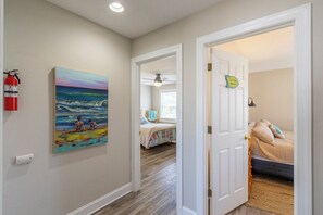 A hallway with a painting of a beach scene, a fire extinguisher, a door leading to a bedroom with a bed and a nightstand, and a ceiling fan. - Pet-friendly oceanside home w/ pool and hot tub by Sun Realty (Kill Devil Hills)