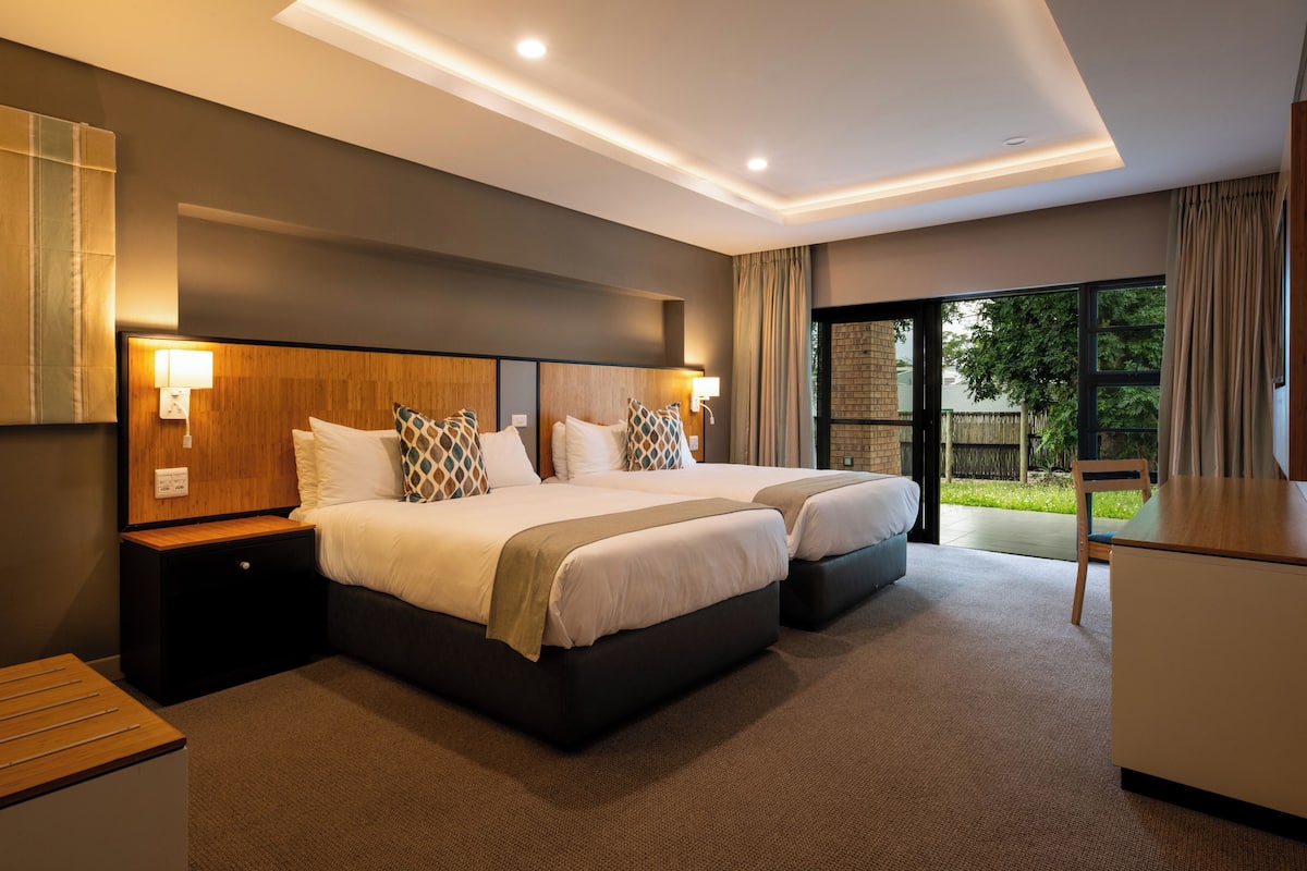 A modern hotel room with two beds, a desk, and a view of the outdoors.