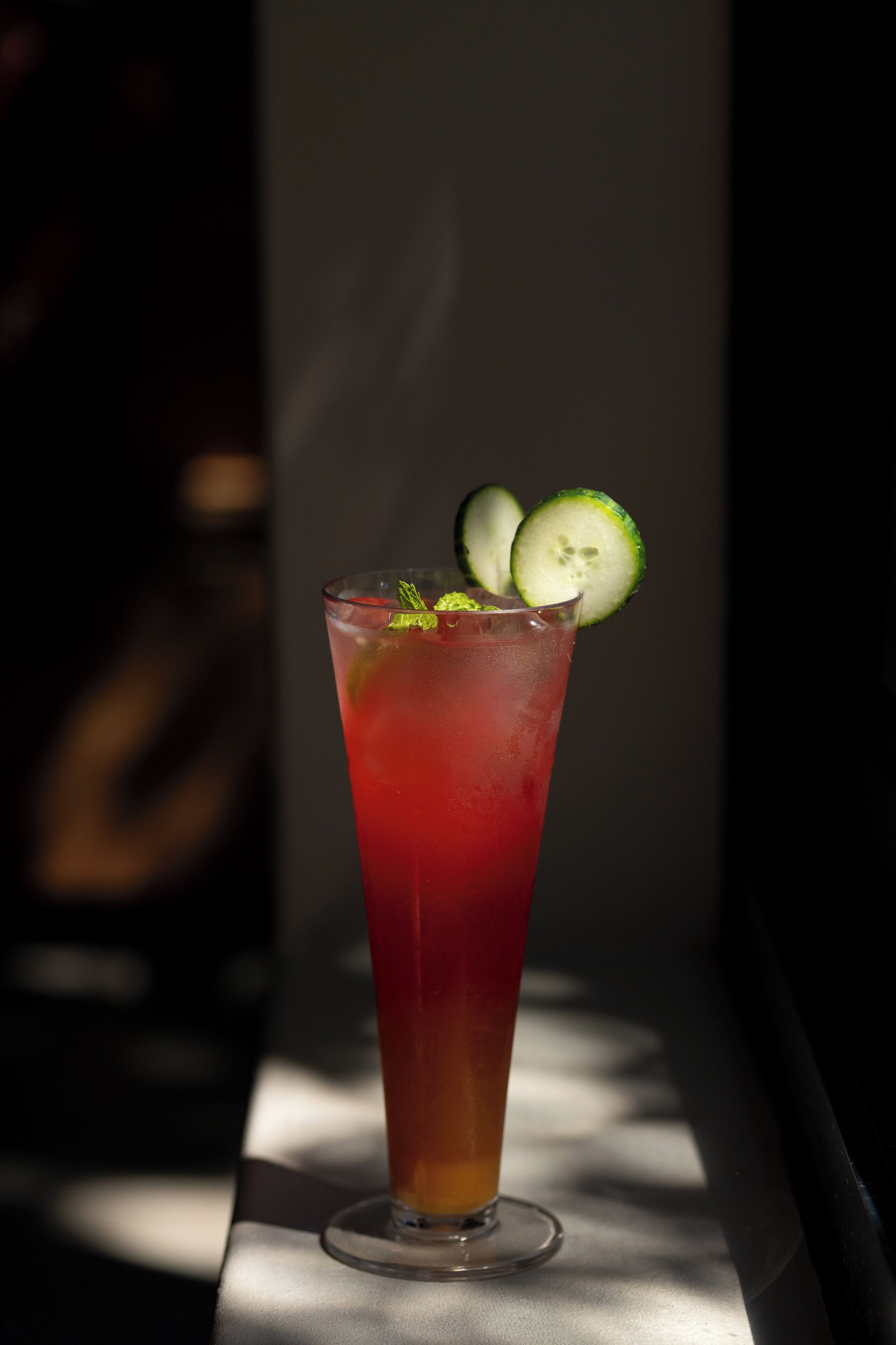 A tall glass with a red beverage, garnished with cucumber slices and mint leaves, placed on a table with a shadow cast on it.