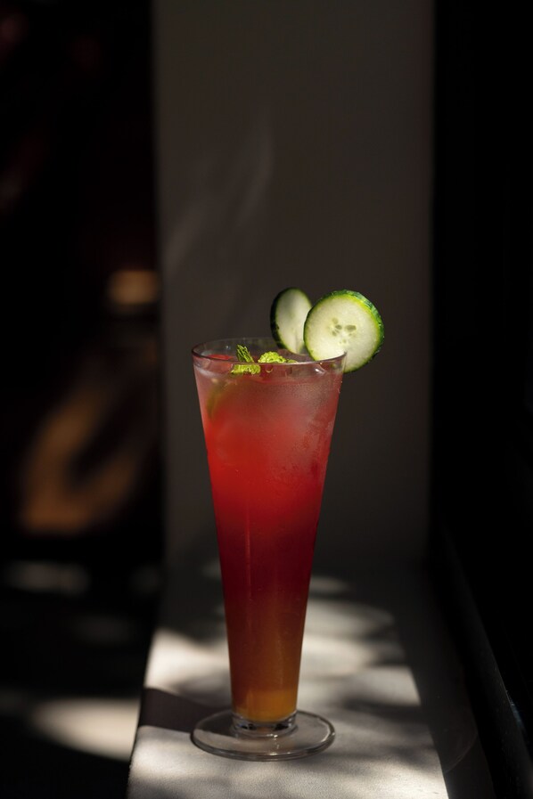 A tall glass with a red beverage, garnished with cucumber slices and mint leaves, placed on a table with a shadow cast on it.