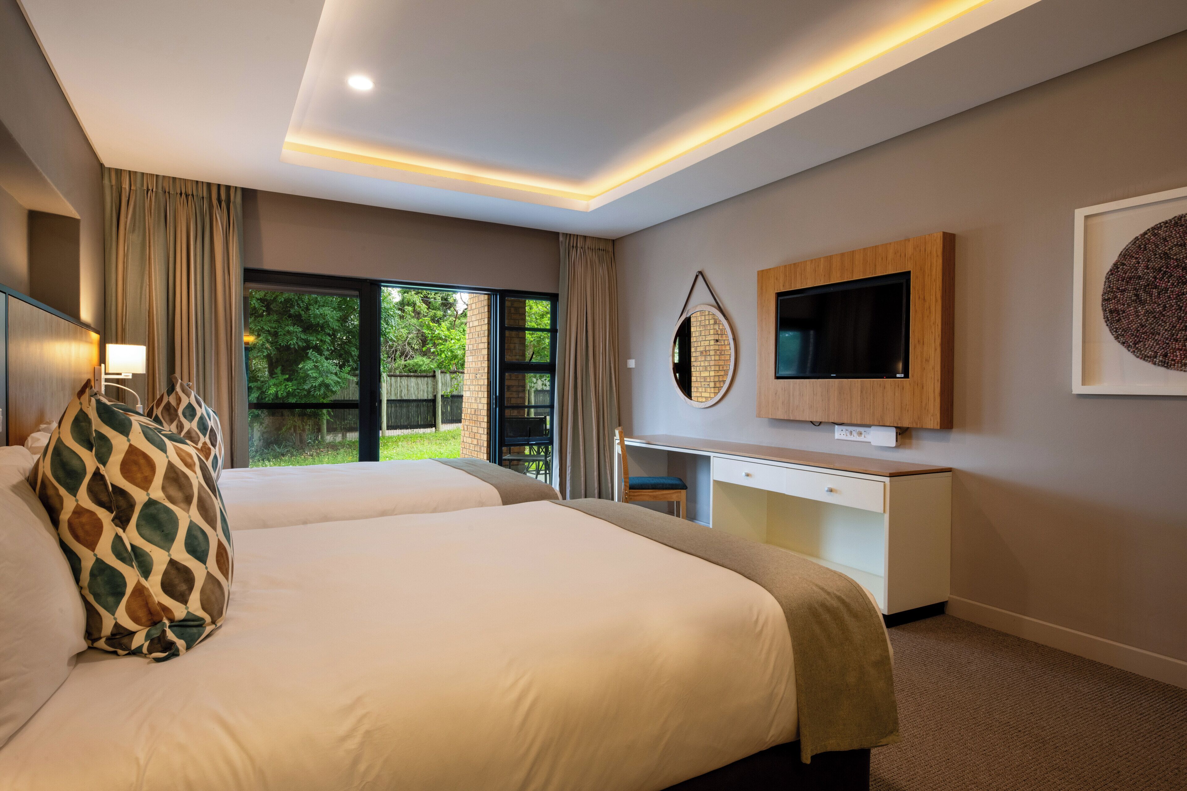 A modern hotel room with two beds, a desk, a TV, and a large window offering a view of greenery.