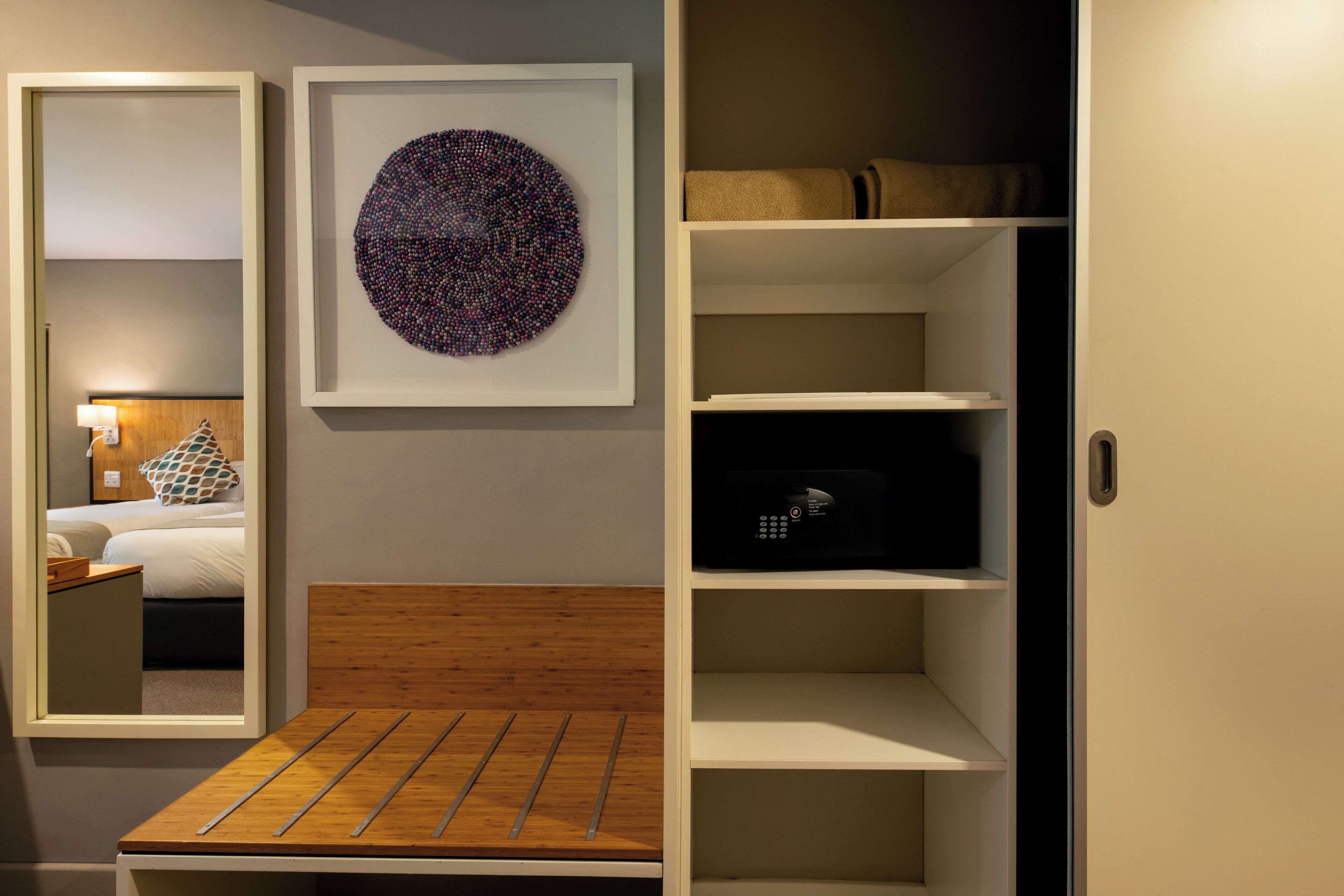 A bedroom with a bed, bedside table, and a mirror. There is a framed artwork on the wall and a shelf with a black object.
