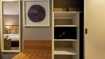 A bedroom with a bed, bedside table, and a mirror. There is a framed artwork on the wall and a shelf with a black object.