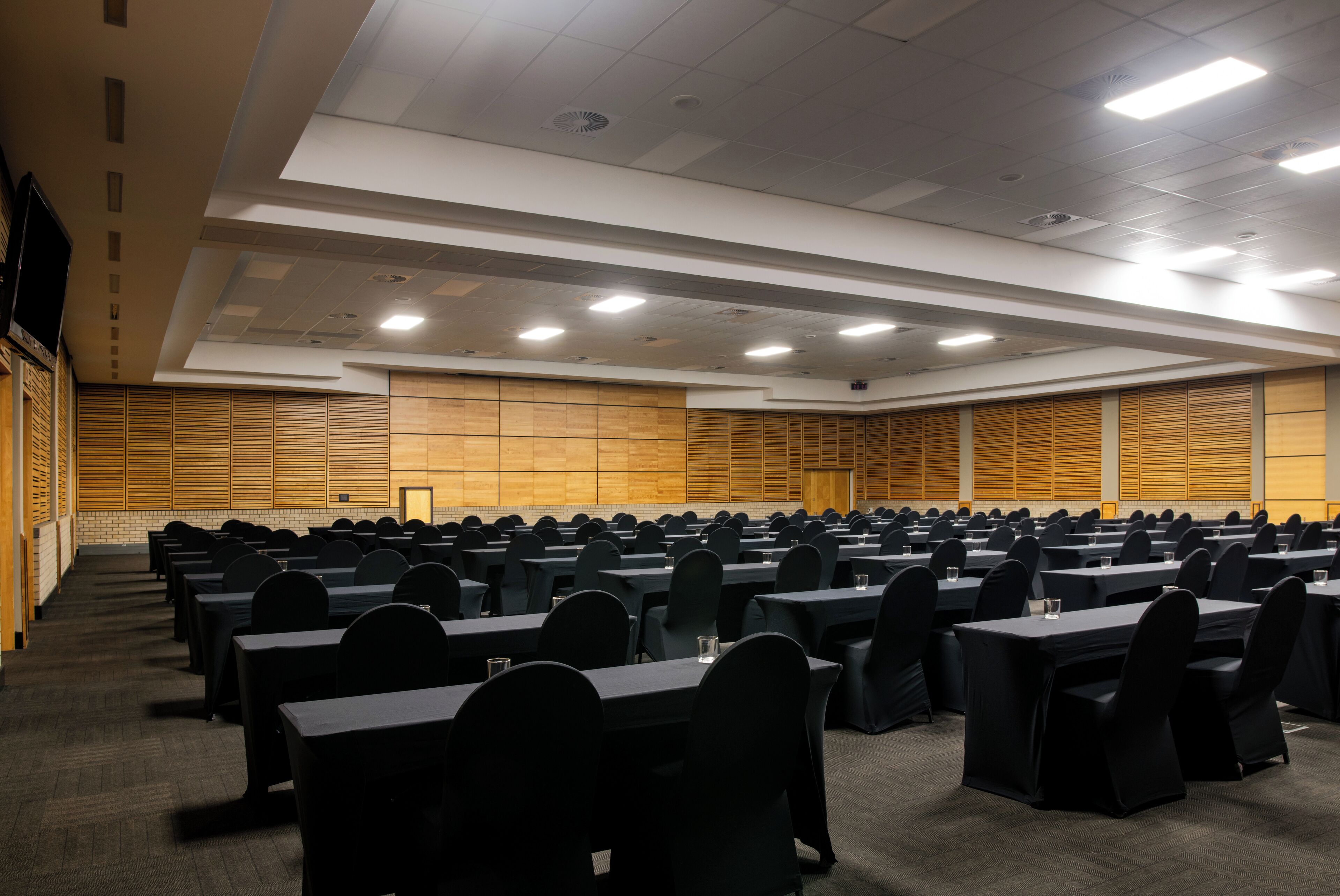 A large conference room with rows of tables and chairs, equipped with ceiling lights and a wooden panel wall.