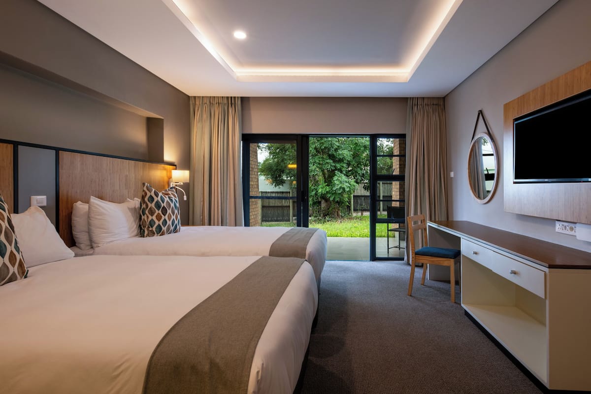 A hotel room with two beds, a desk, a chair, a TV, and a large window with a view of greenery.