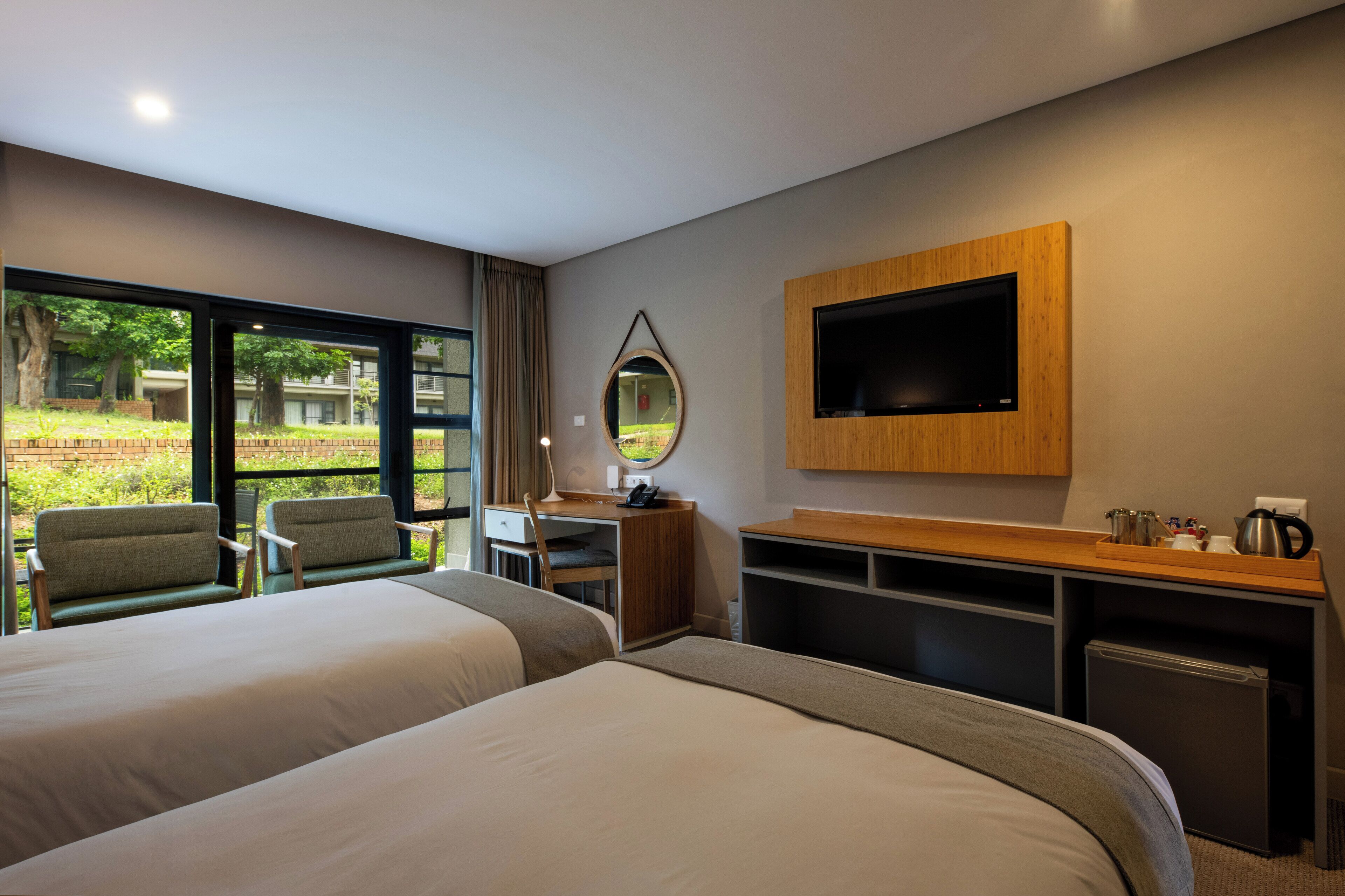 A hotel room with two beds, a desk, a television, and a view of greenery outside.
