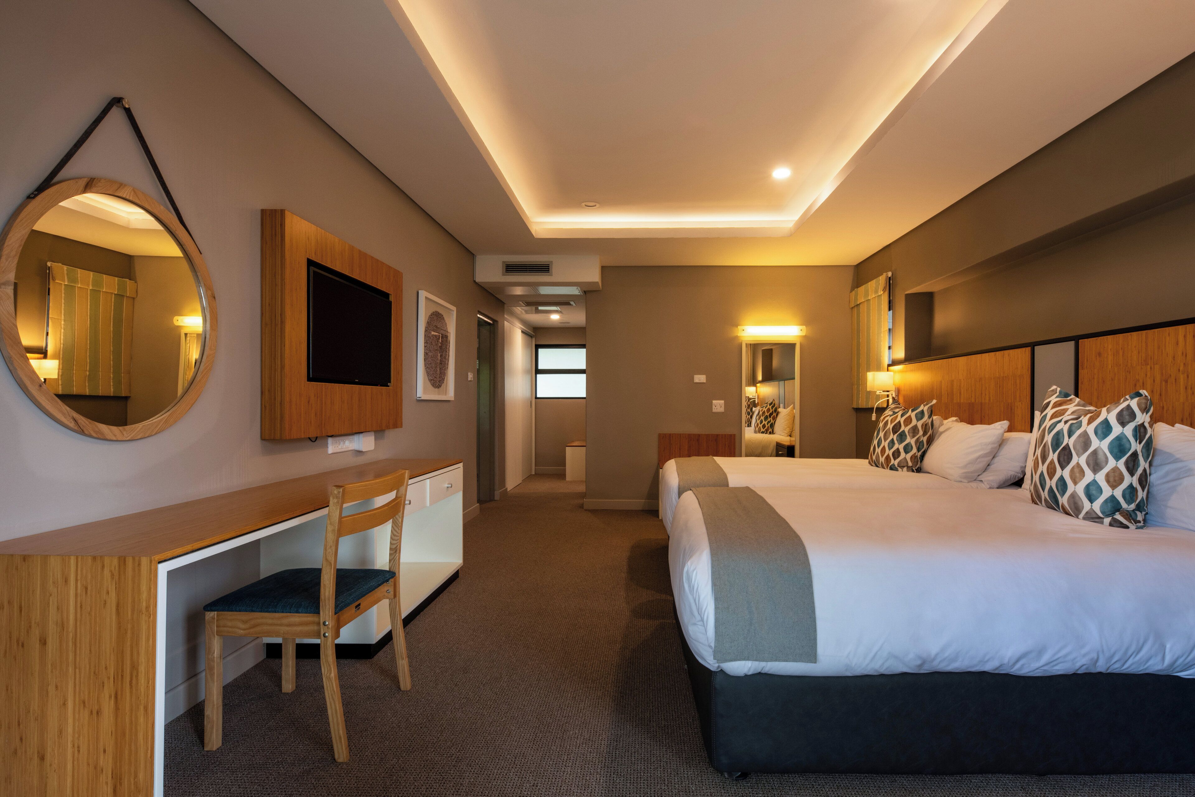Une chambre d’hôtel équipée d’un bureau, de deux lits, d’un grand miroir et d’une télévision.