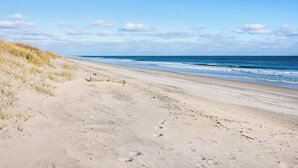 Una playa arenosa con huellas que se adentran en el agua, cielo despejado y dunas al fondo.