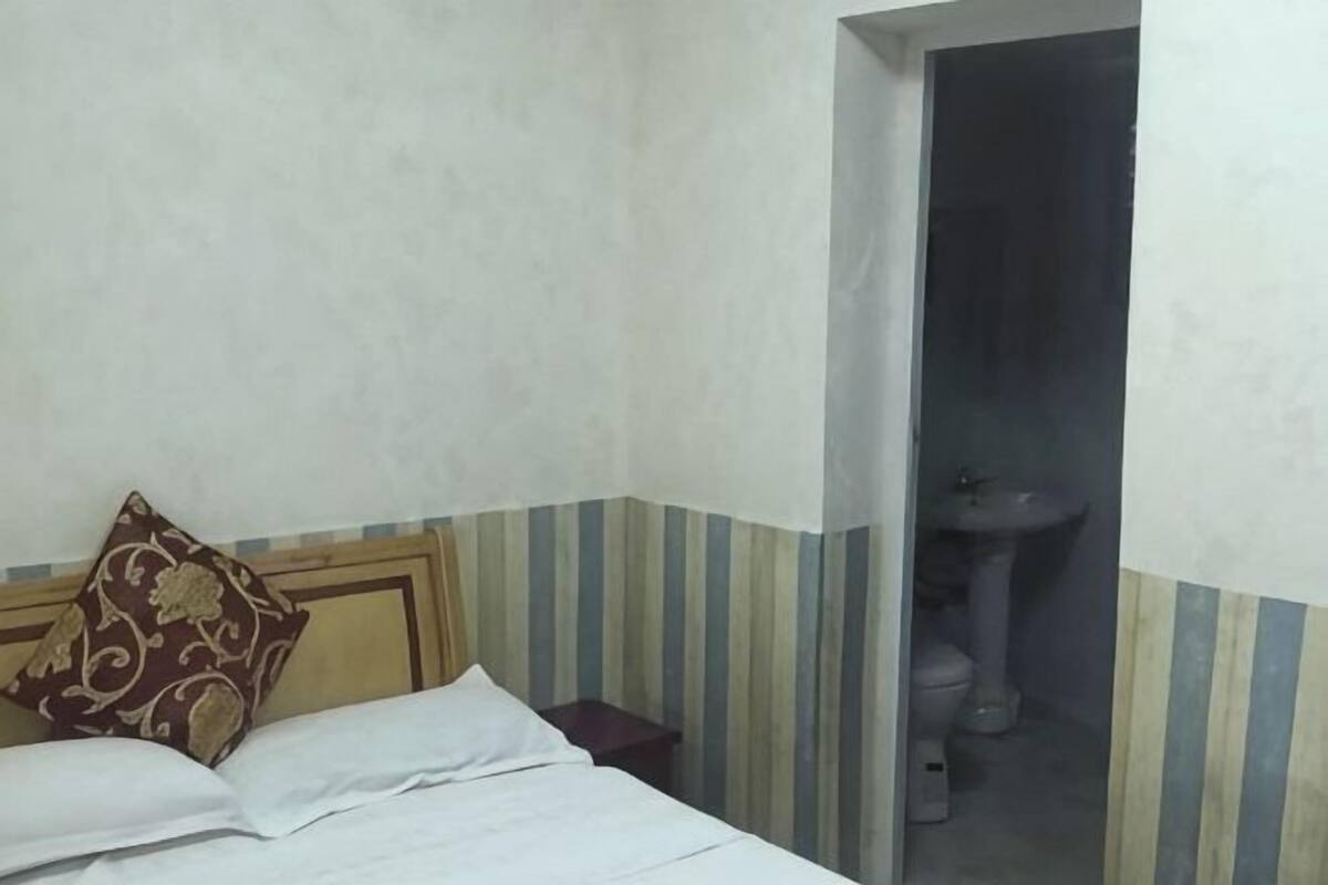 A bedroom with a bed, pillows, a bedside table, and a bathroom with a sink and toilet.