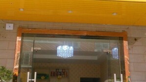 A hotel entrance with a glass door, a chandelier, and a visible interior with a tiled wall and a shelf with items.