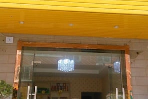 A hotel entrance with a glass door, a chandelier, and a visible interior with a tiled wall and a shelf with items.