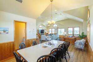 A spacious dining area with a large table and chairs, a kitchen in the background, and a view of the outdoors through large windows.