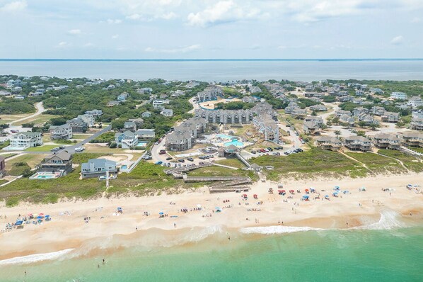 An aerial view of a coastal residential area with houses, a beach, and a large building complex with a swimming pool.