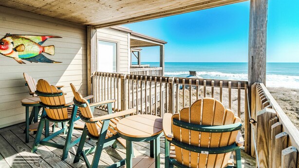 Une terrasse en bois avec des fauteuils Adirondack et une table, donnant sur une plage et l’océan.