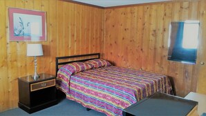 Une chambre avec un mur lambrissé en bois, un lit recouvert d’une couverture à rayures colorées, une table de chevet avec une lampe et un tableau encadré accroché au mur.