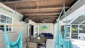 Een overdekt terras met een houten plafond, een eettafel met stoelen, een blauwe bank en twee hangmatten.