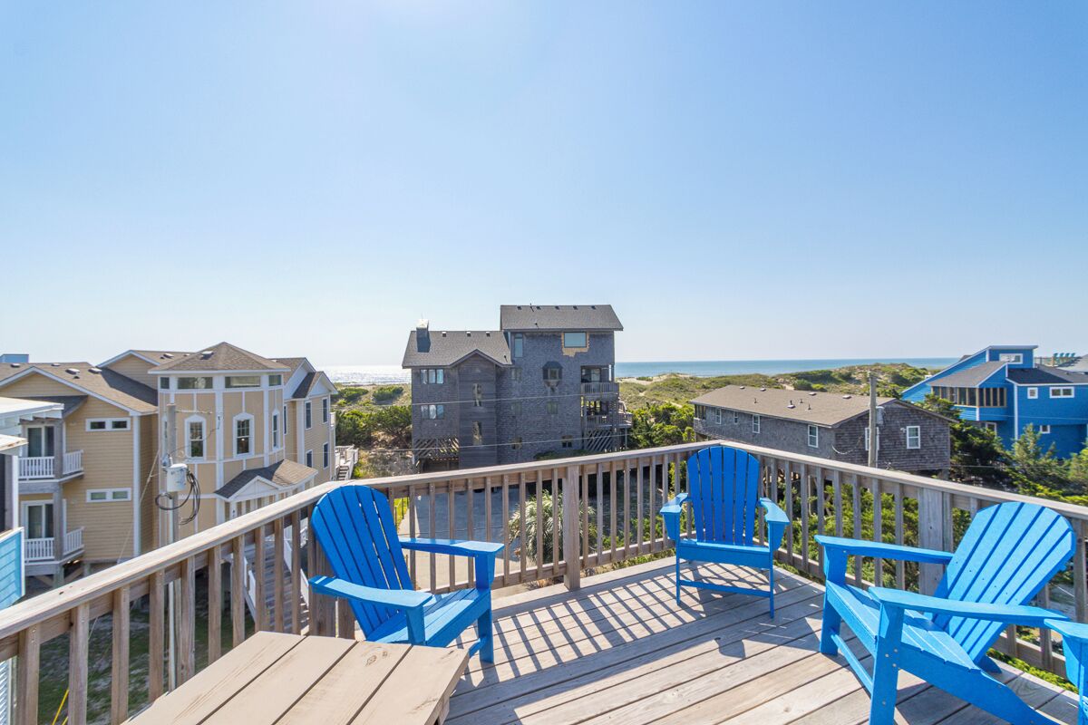Una terraza de madera con sillas Adirondack azules que da a una zona costera con casas y un cielo despejado.