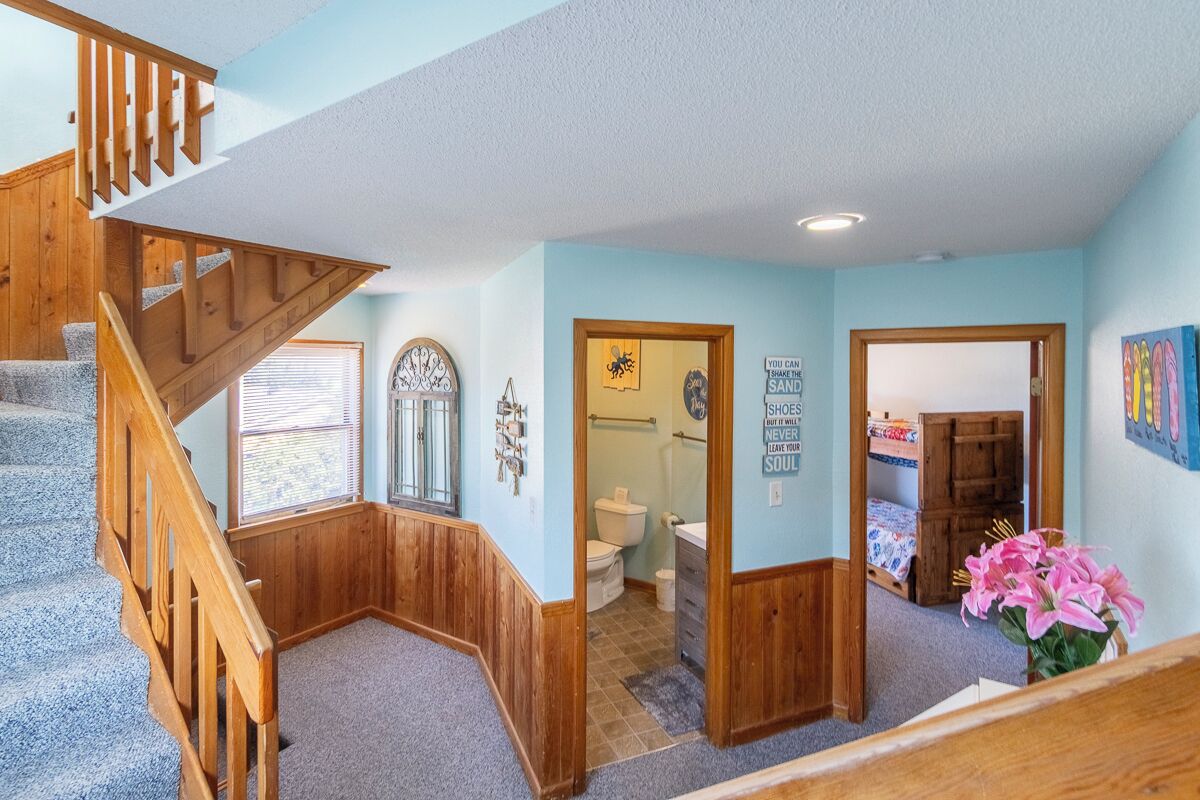 Interior de una casa de varios niveles con una escalera de madera, un baño con inodoro y lavamanos, y un dormitorio con cama y cómoda.