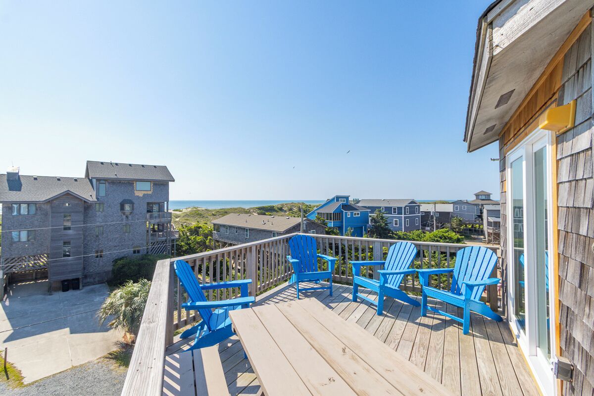 Une terrasse en bois avec des chaises bleues, une vue sur l’océan et plusieurs bâtiments en arrière-plan.