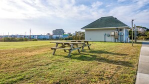 Un área de picnic con una mesa y bancos de madera, un pequeño edificio con techo verde, y cielo despejado.