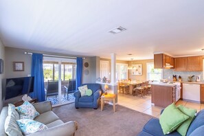 A living room with a sofa, a blue armchair, a TV, and a dining area with a kitchen in the background.