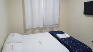 A single bed with a white mattress and a blue polka dot bedspread. There is a white curtain on the window, and an air conditioning unit is mounted on the wall. - Hotel londres (Gama)