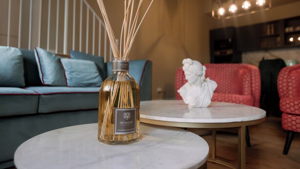 Ein Zimmer mit einem runden Marmortisch, einer Flasche Dr. Ravanes Fragrance Collection, einem blauen Sofa mit roter Ziernaht und einem rot gemusterten Sessel.