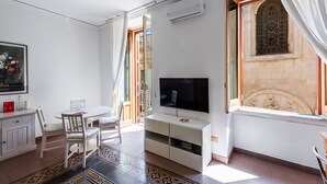 A modern living room with a flat-screen TV, a dining table with chairs, a patterned rug, and a decorative window with stained glass.