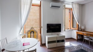 A modern living room with a round dining table, white furniture, a flat-screen TV, and a balcony with a wrought iron gate.