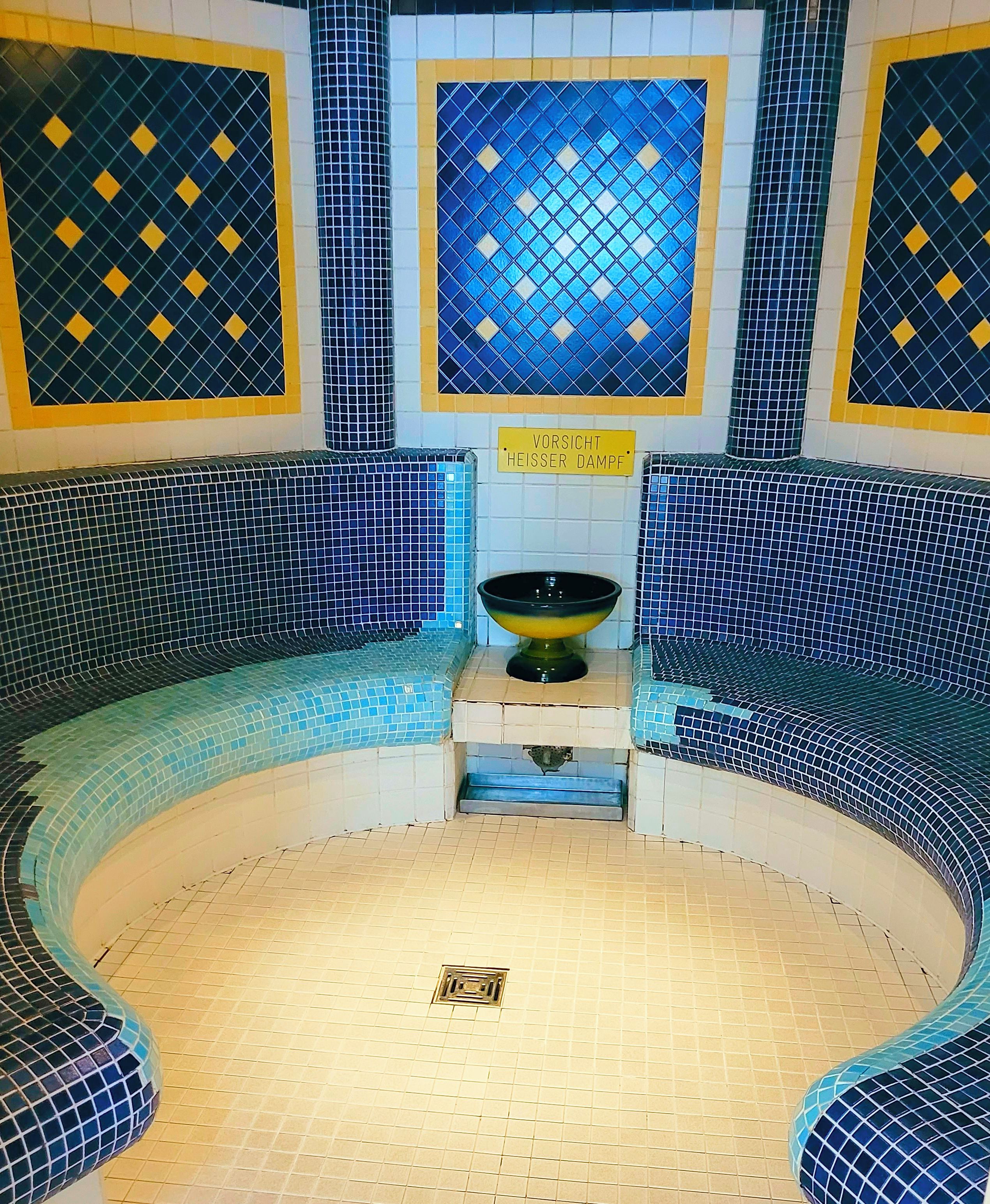 Een sauna met een centrale bank, een schaal en een bordje met de tekst 'VORSICHT HEISSER DAMPF'.