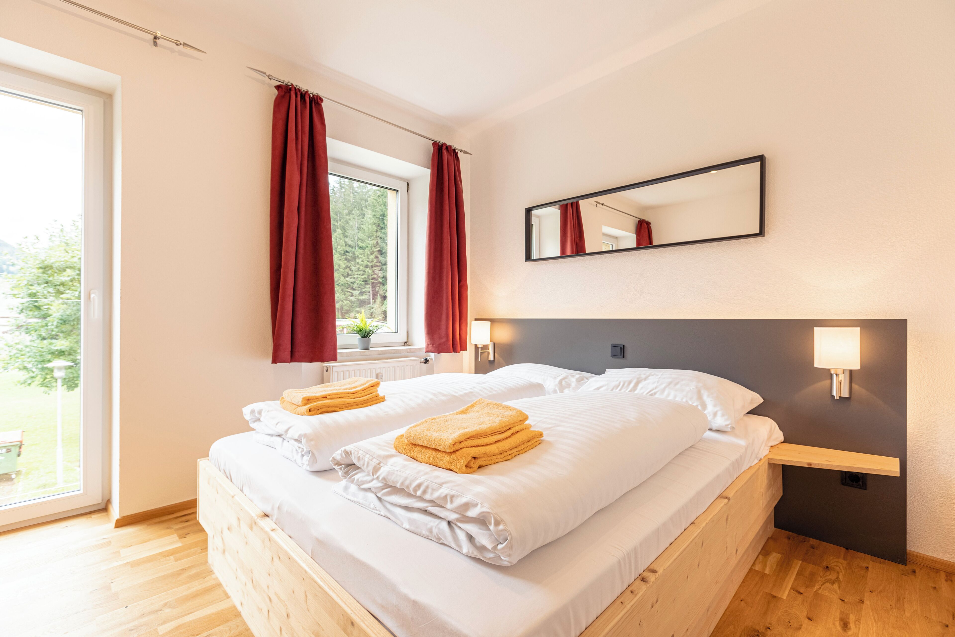 Ein Doppelbett mit weißen Leinentüchern und zwei Handtüchern, ein Nachttisch mit Lampe, ein Fenster mit roten Vorhängen und ein Spiegel an der Wand.