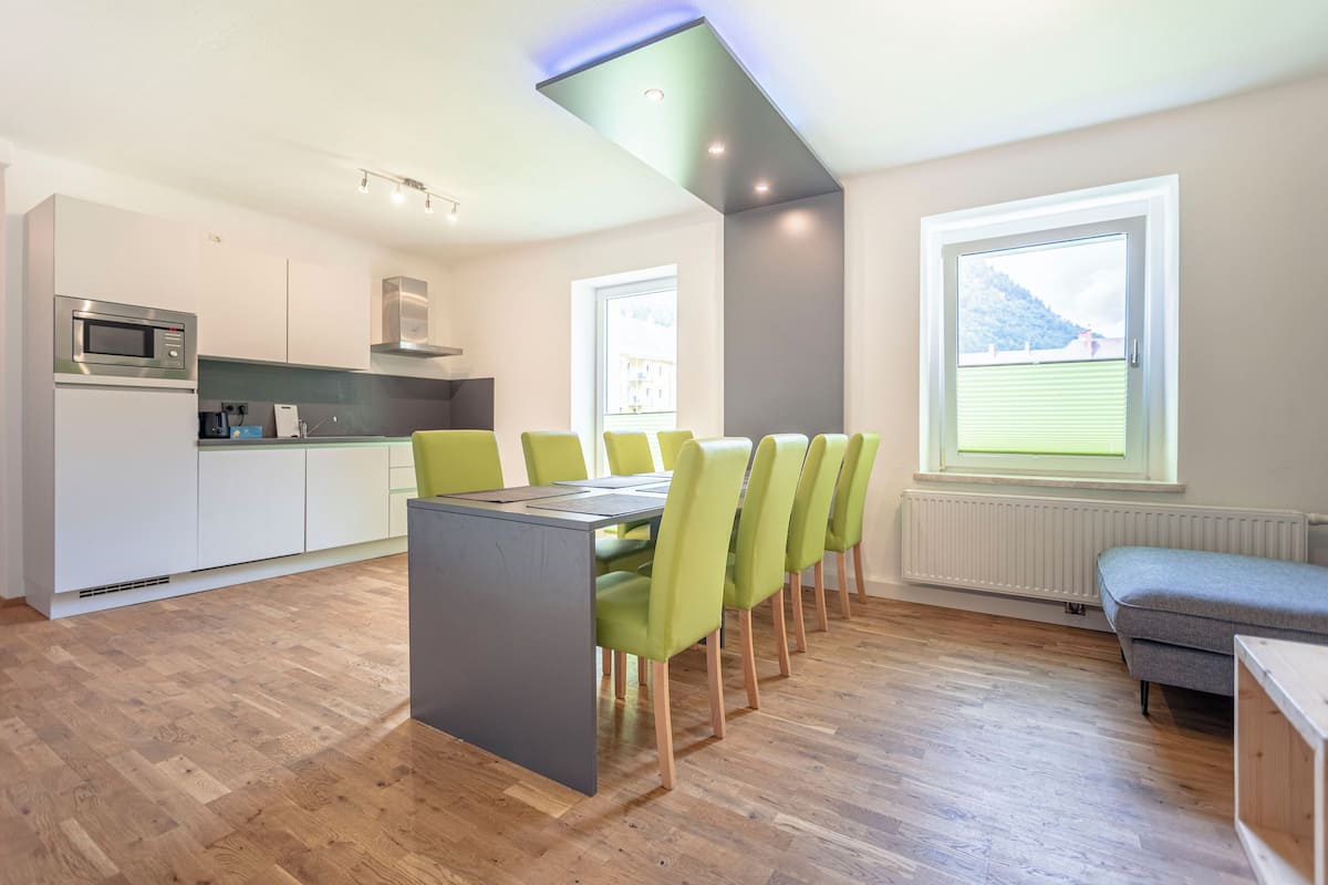 Una cocina moderna con microondas integrado, un comedor con mesa gris y sillas verdes, y una ventana con vistas a una montaña.