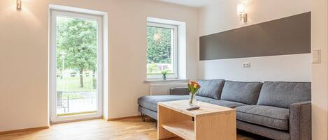A modern living room with a grey sofa, wooden coffee table, and a view of a green garden through a large window.