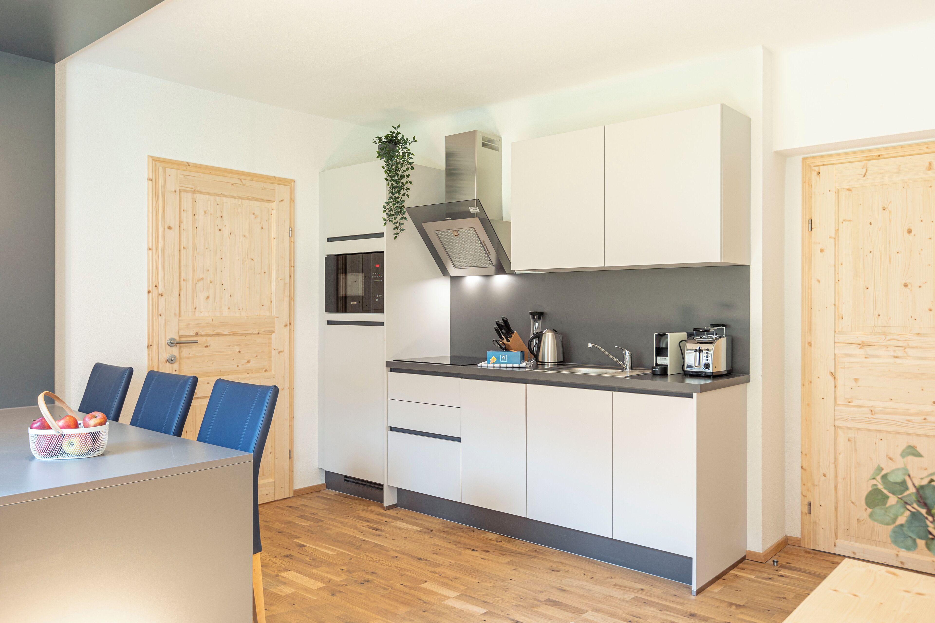 A modern kitchen with white cabinets, a black countertop, and a built-in oven. There is a dining area with blue chairs and a table, and a wooden door on the left.