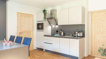 A modern kitchen with white cabinets, a black countertop, and a built-in oven. There is a dining area with blue chairs and a table, and a wooden door on the left.