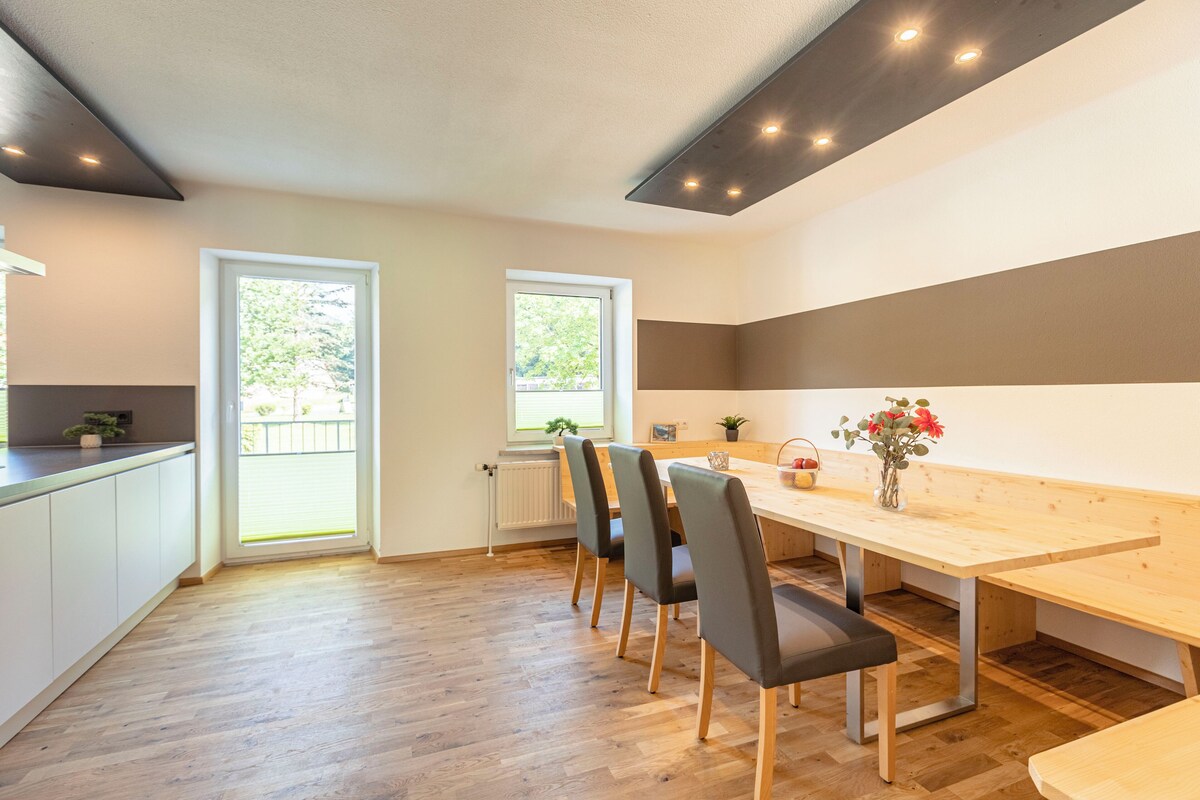 Una cocina moderna con armarios blancos, una mesa de comedor de madera y un banco. Hay dos ventanas que proporcionan luz natural y una planta en maceta sobre la encimera.