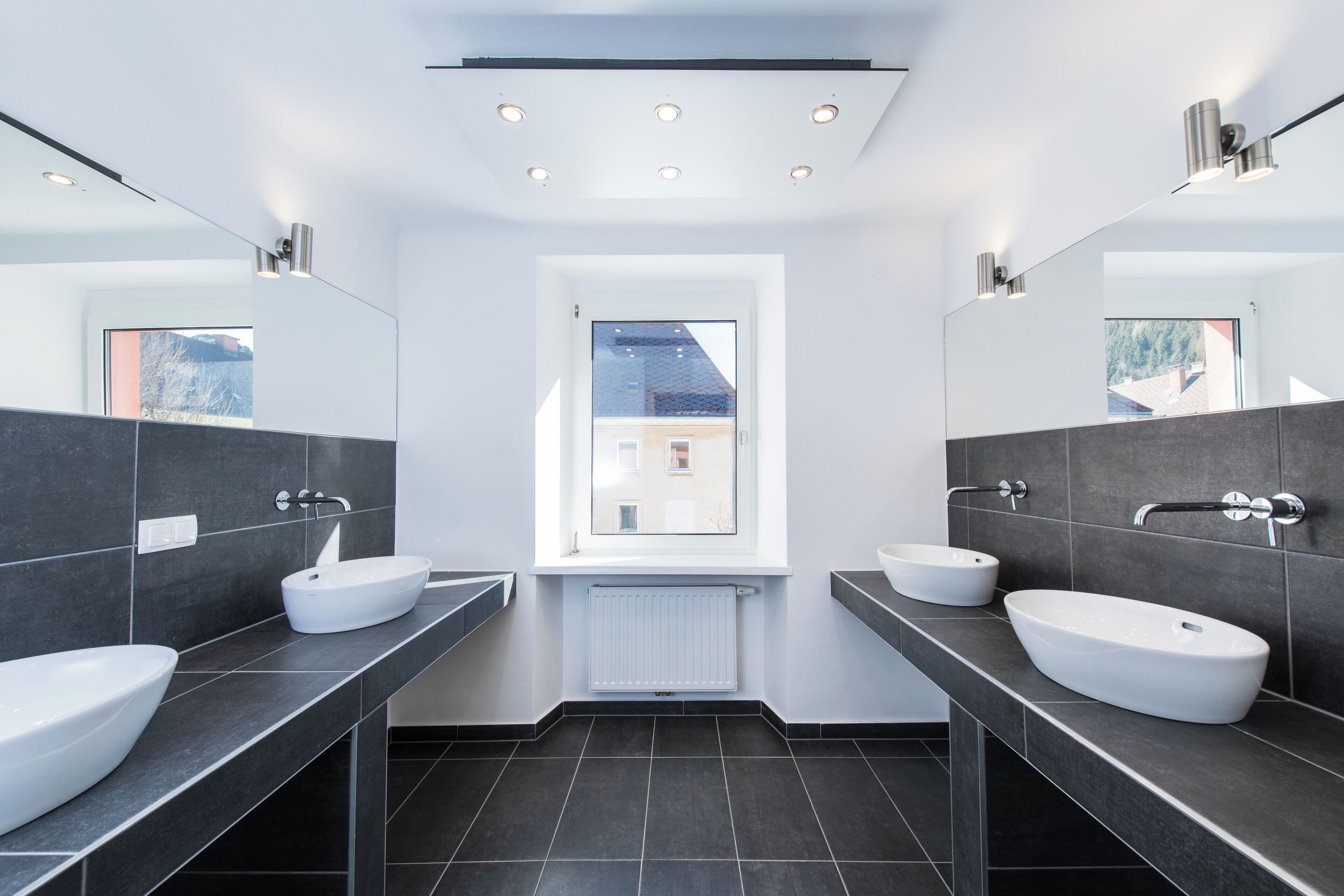 A modern bathroom with two white sinks, dark tiled flooring, and a window providing a view of a building outside.
