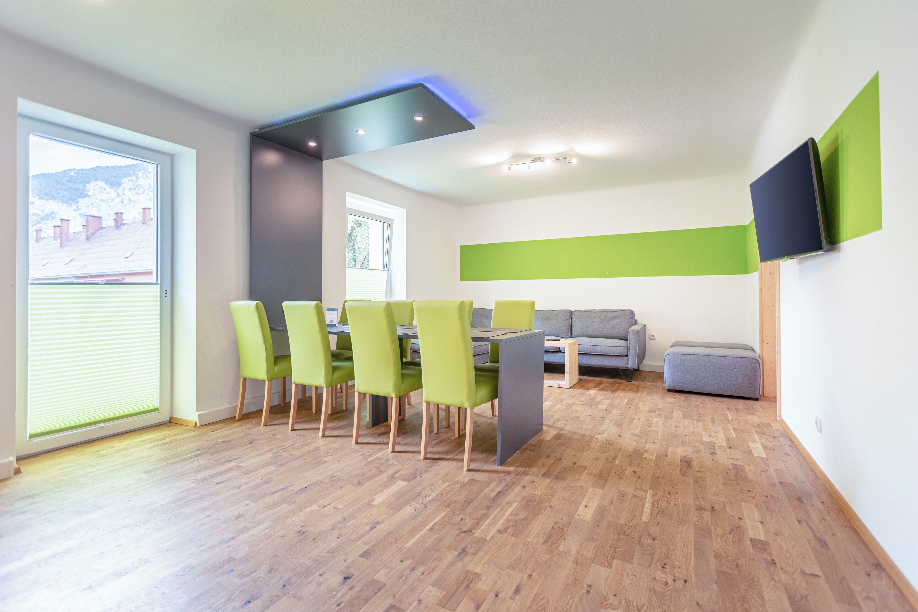 A modern dining area with a wooden floor, green chairs, a grey table, and a grey sofa.