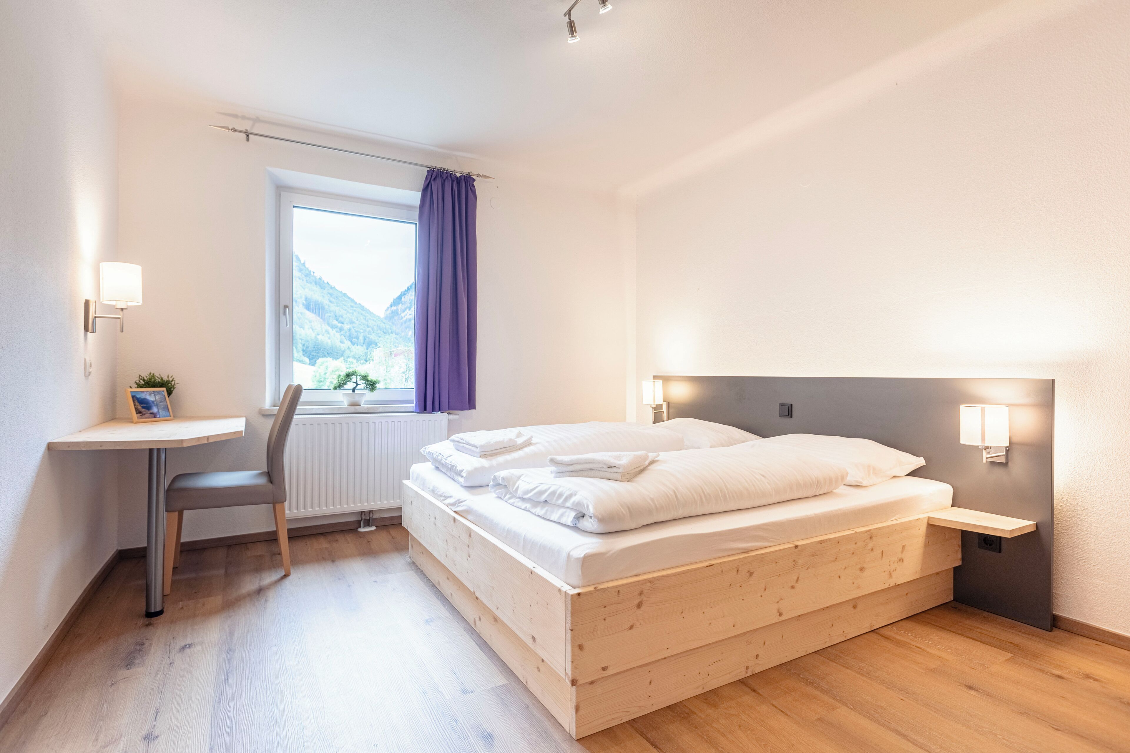 A bedroom with a wooden bed, a desk, and a window with a view of a mountain.