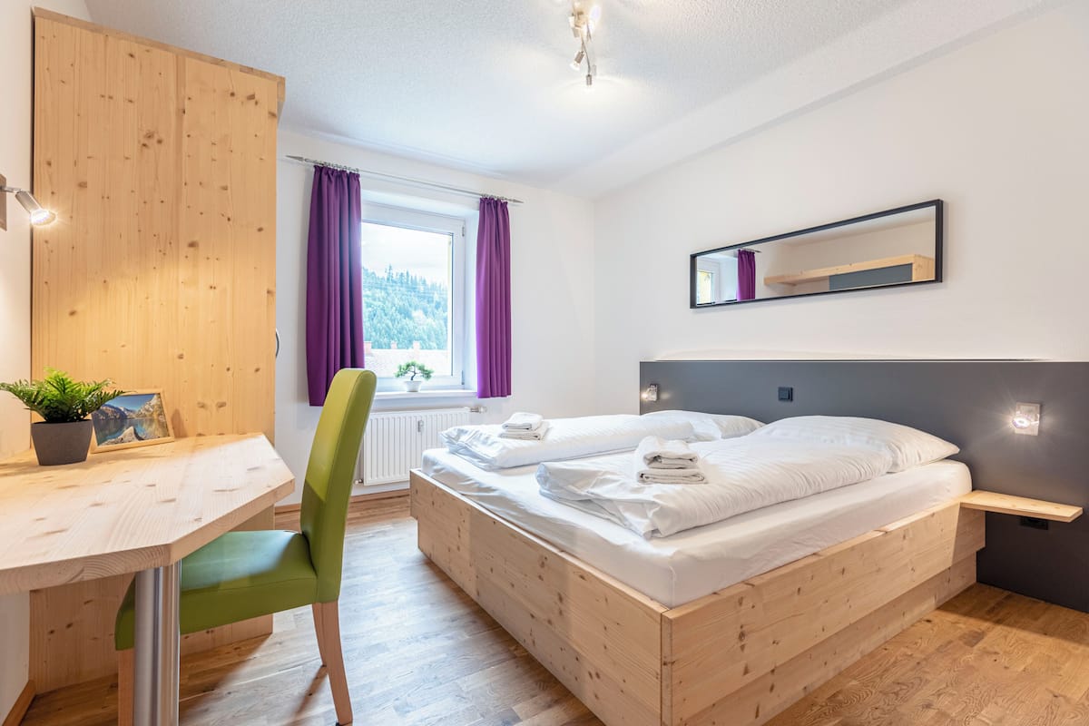 Un dormitorio con una cama de madera, un escritorio, una silla, una ventana con cortinas moradas y un espejo en la pared.