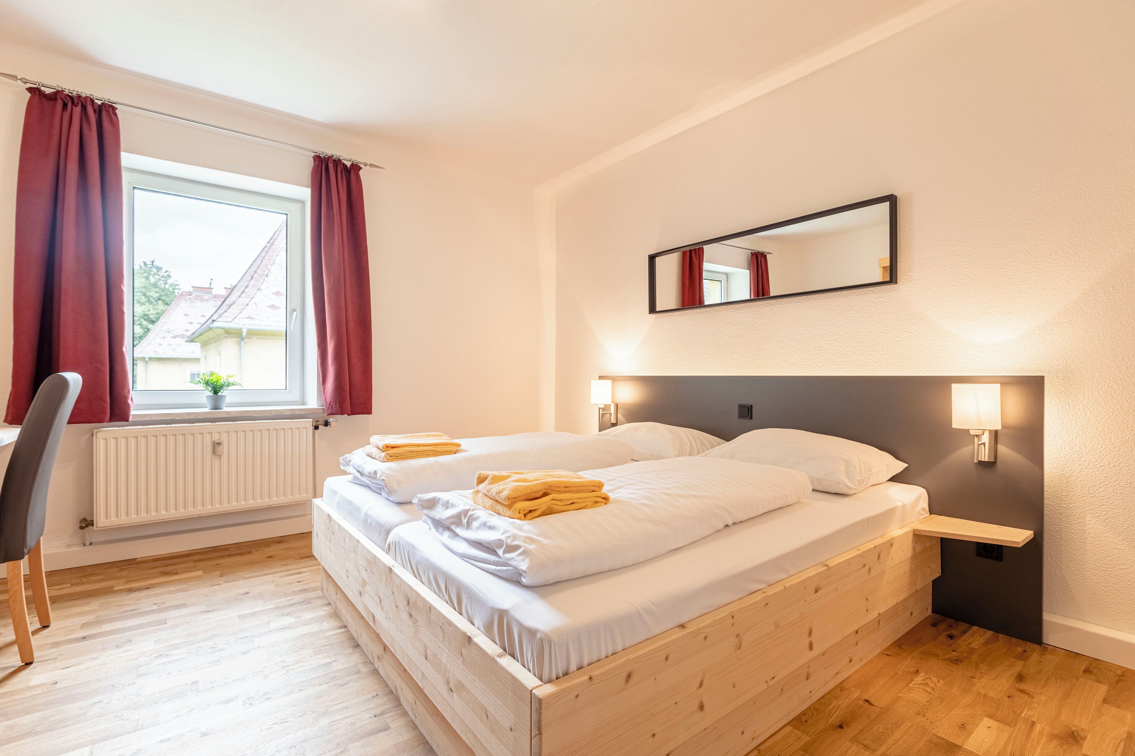 A double bed with white linens and a wooden headboard, two bedside tables with lamps, a window with red curtains, and a radiator.