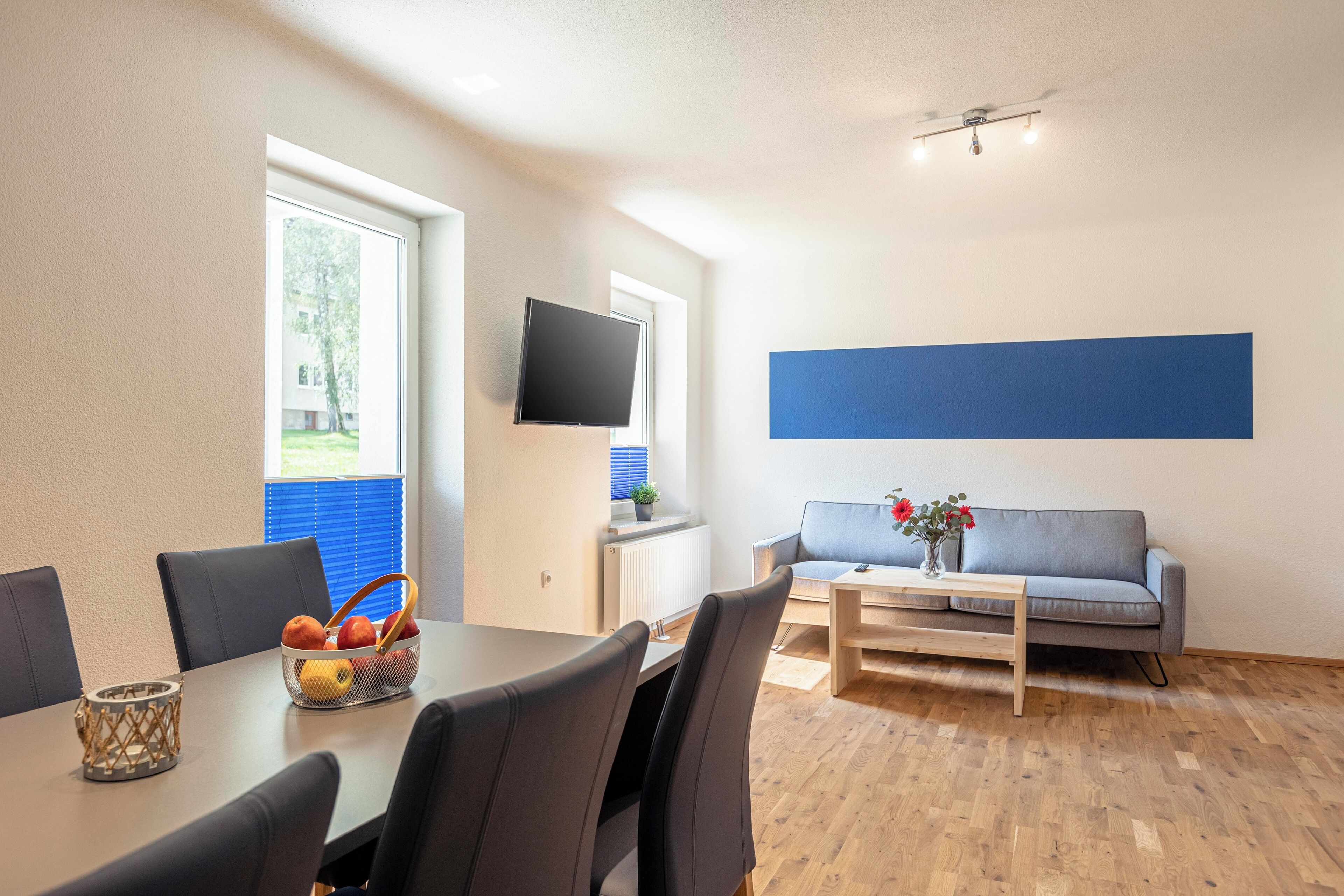 A modern living room with a dining area, a grey sofa, a flat-screen TV, and a large blue accent wall.