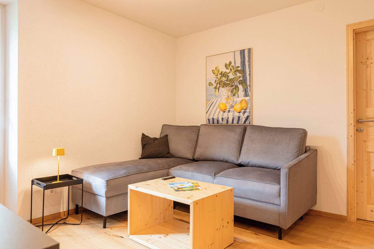Una sala de estar con un sofá gris, una pequeña mesa de centro de madera, un cuadro en la pared y una puerta a la derecha.