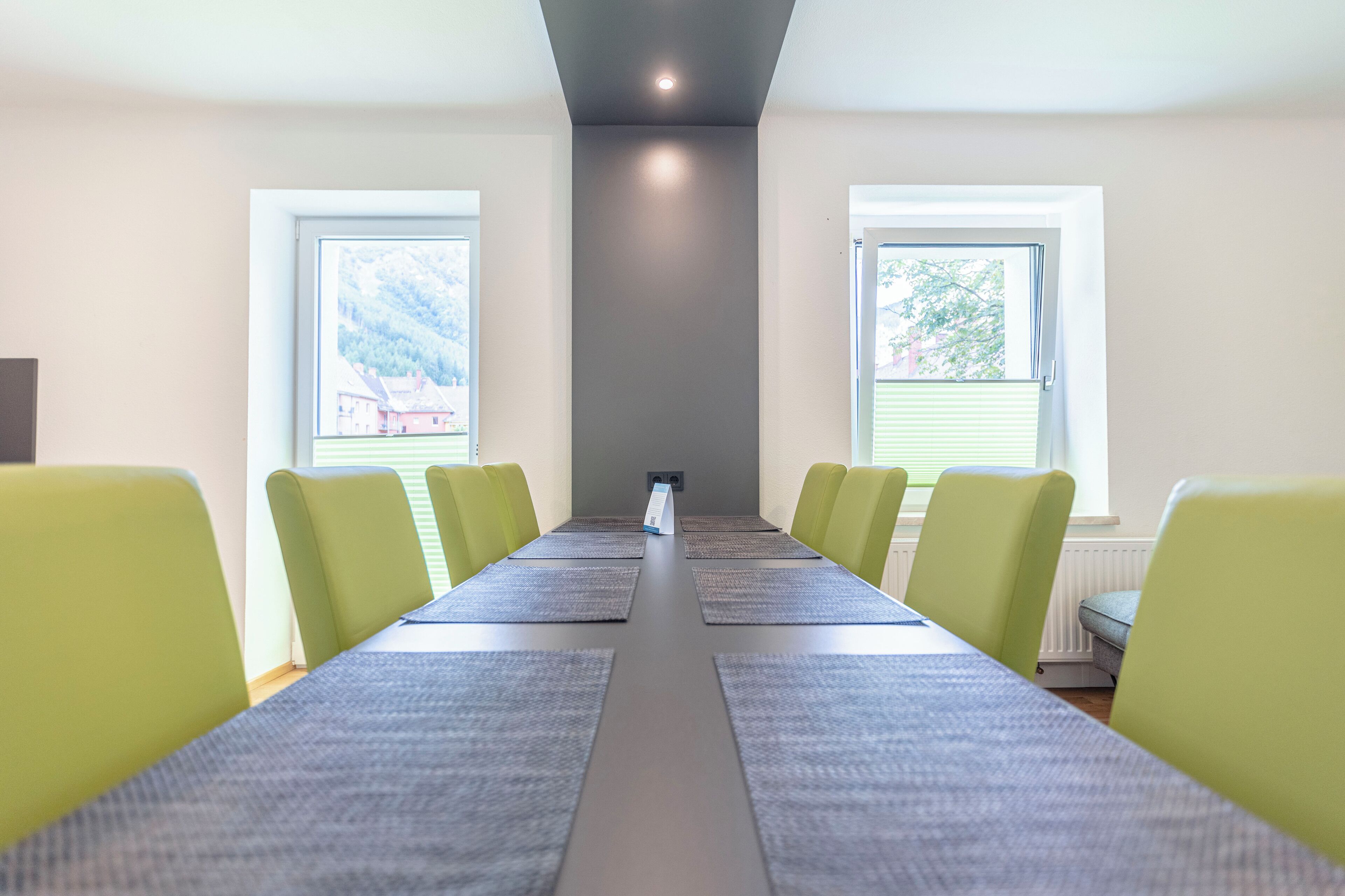 A conference room with a long table, green chairs, and two windows with blinds.
