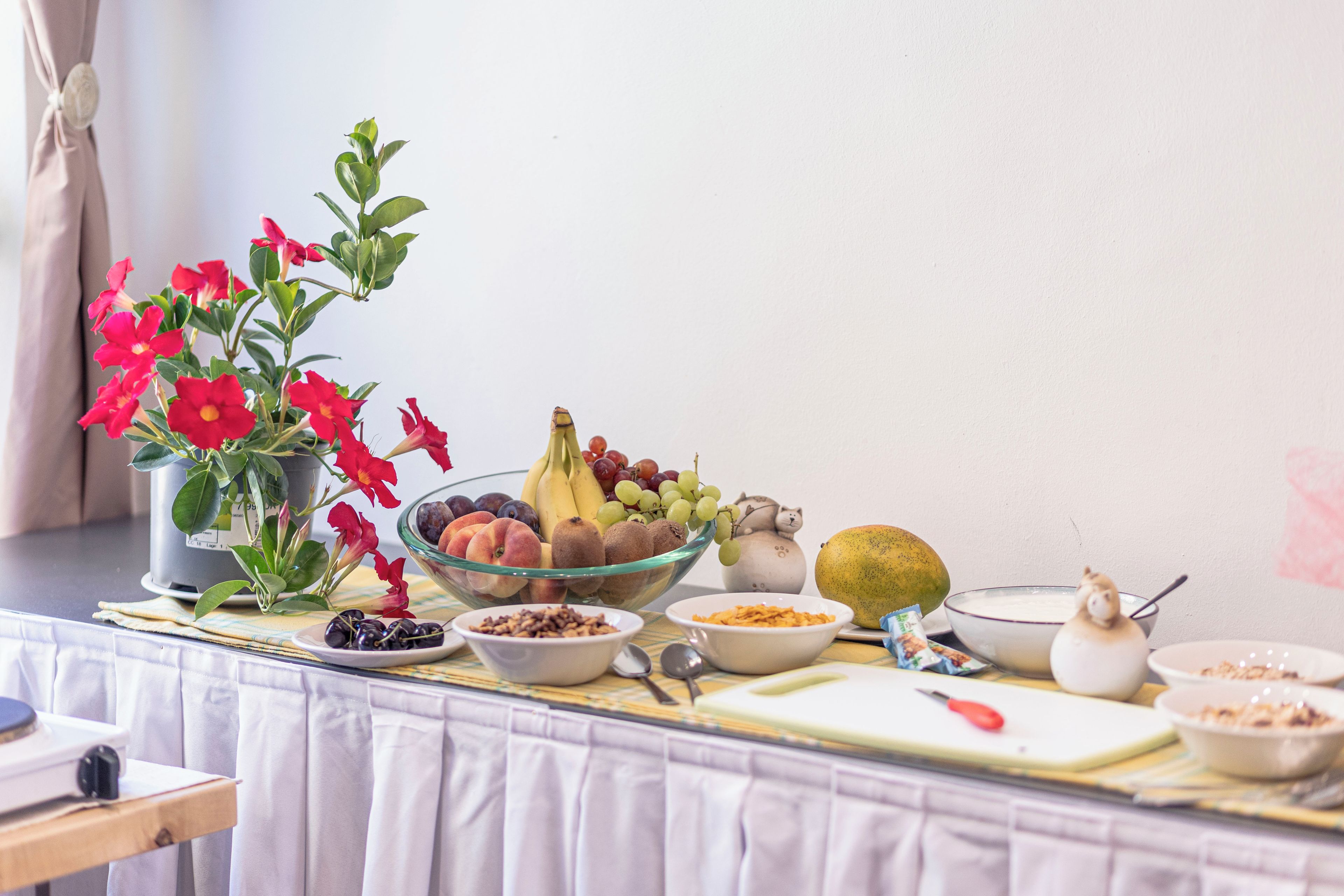 Een tafel met diverse etenswaren, een fruitschaal en een potplant met rode bloemen.