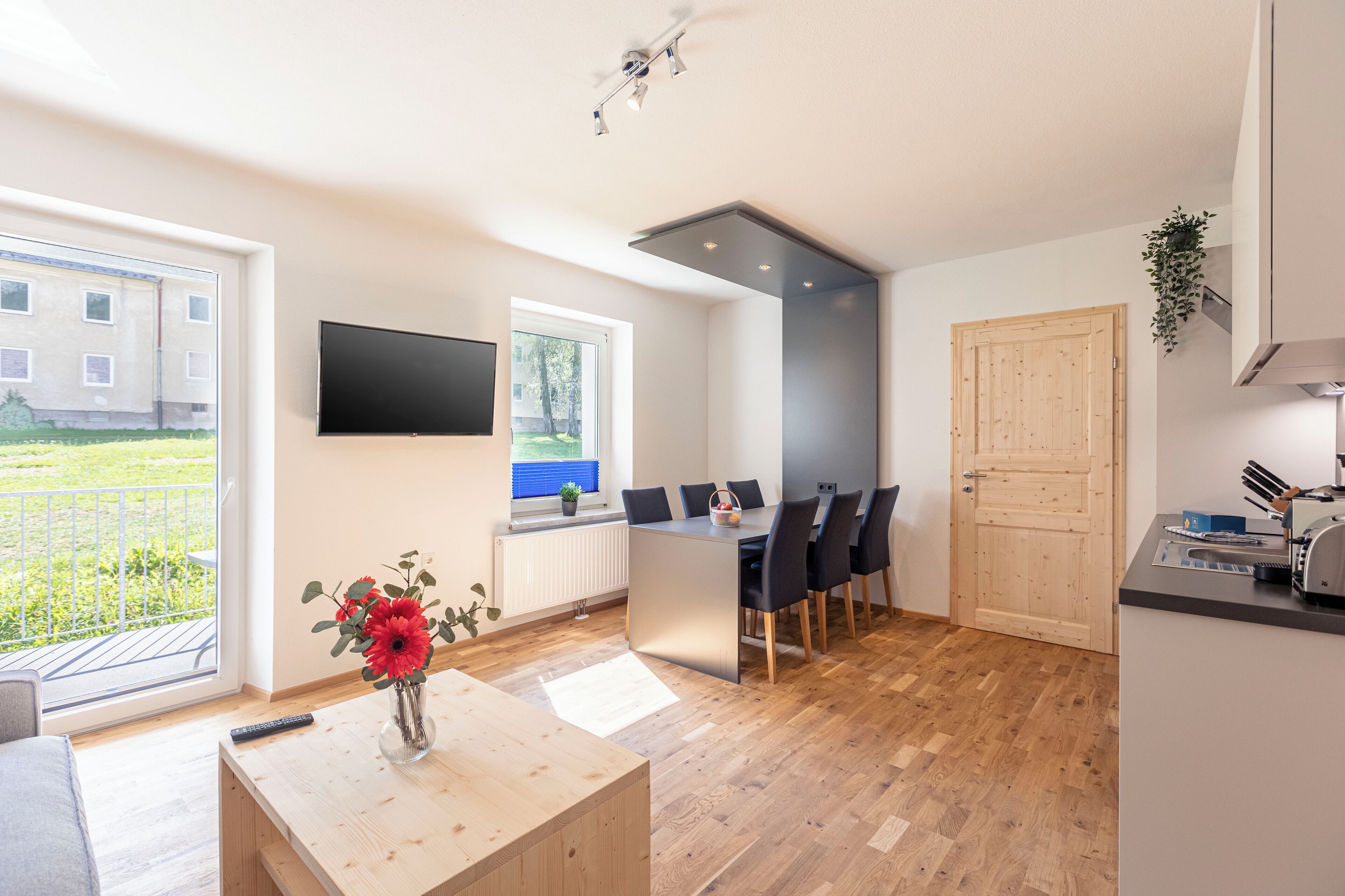 A modern kitchen with a dining area, a TV mounted on the wall, and a view of the outside.
