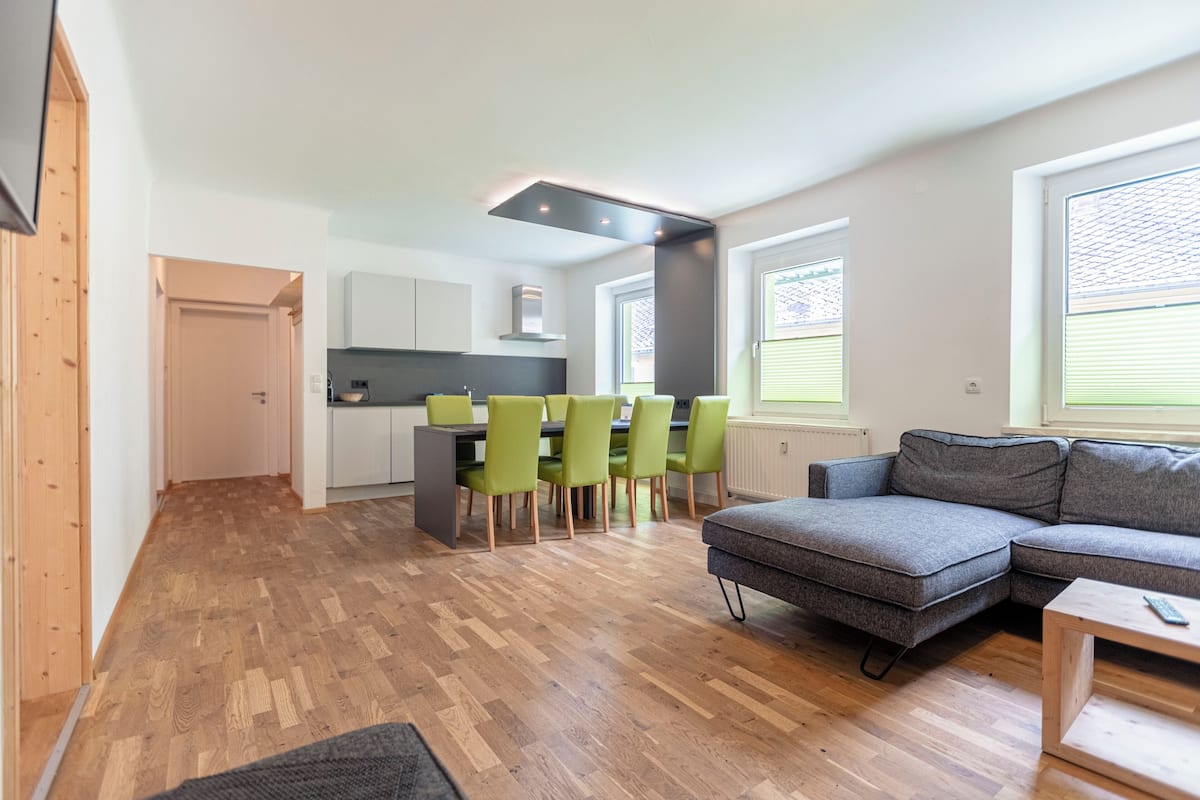 Una sala de estar moderna con un sofá seccional gris, una mesa de centro de madera y un comedor con sillas verdes y una mesa gris.