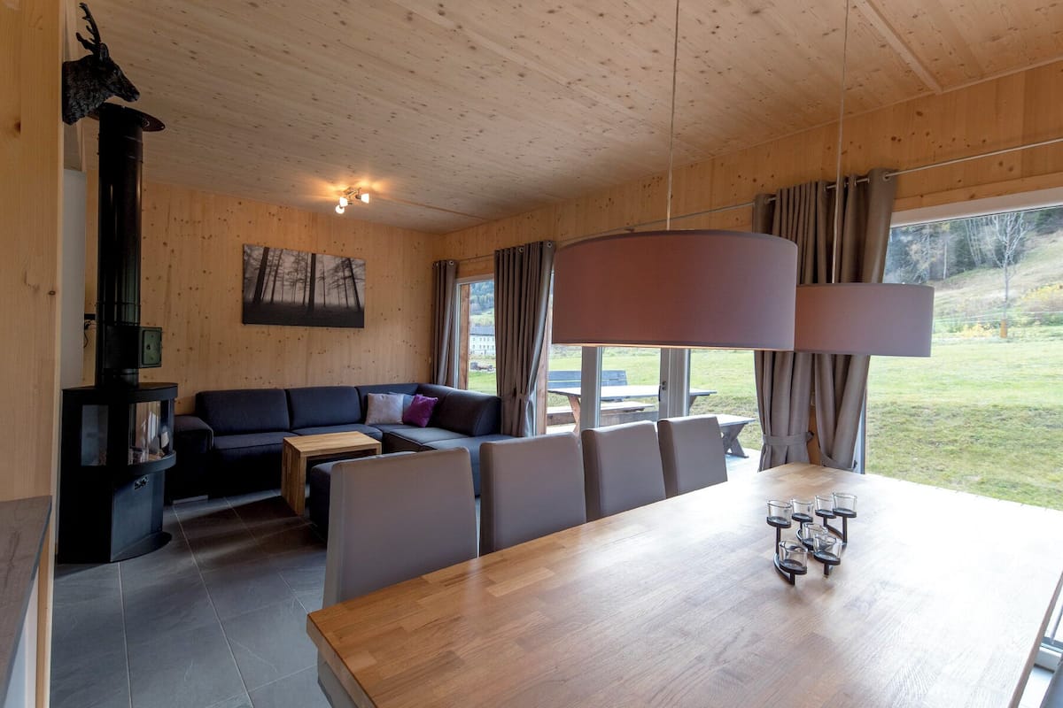 Interior acogedor de una cabaña con techo de madera, un sofá, una mesa de comedor y un ventanal con cortinas.