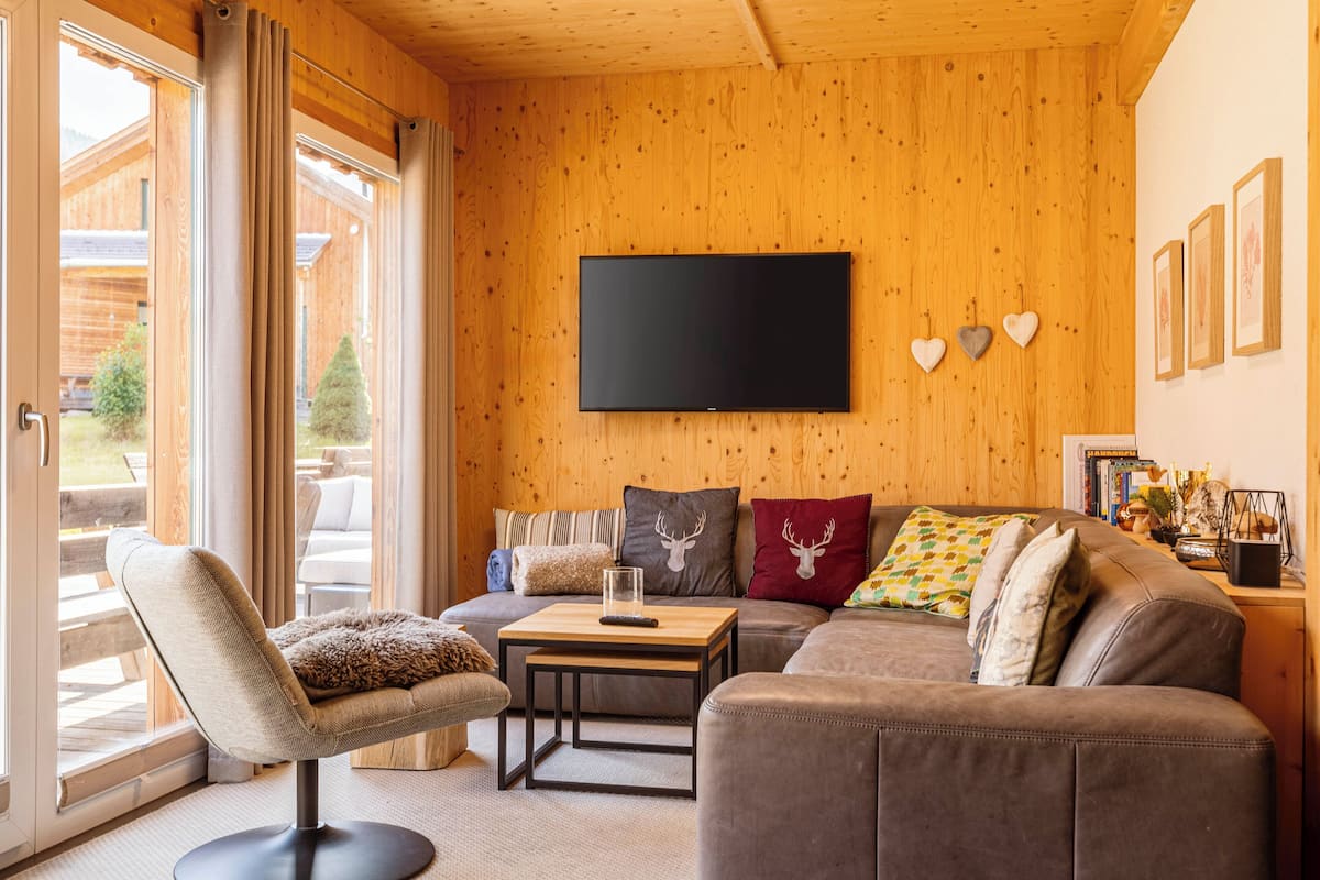 A cozy living room with a wooden interior, a flat-screen TV mounted on the wall, a sofa with various cushions, a small table, and a chair.